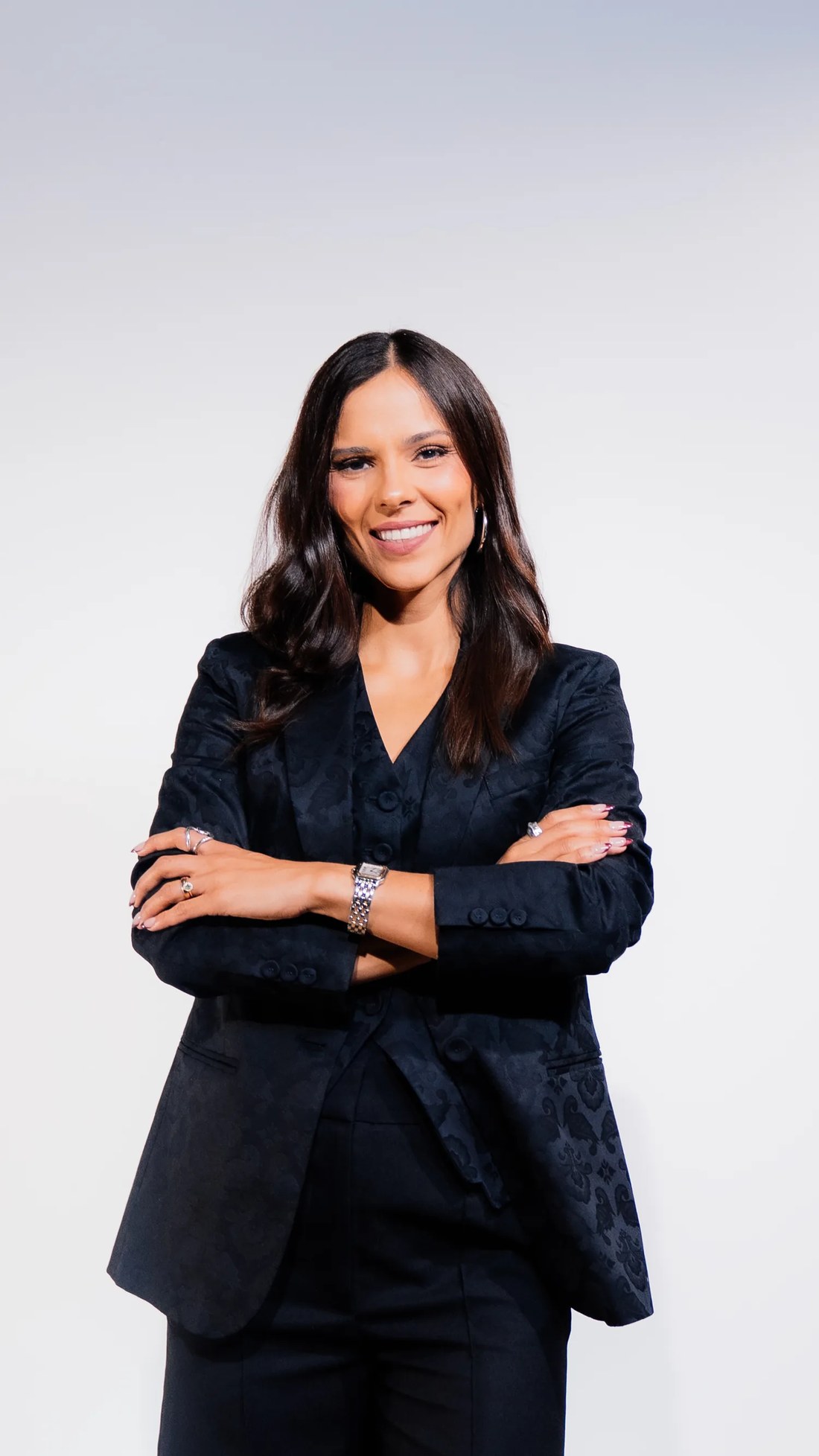 Ana Fidalgo, de traje negro y brazos cruzados, sonriendo a cámara sobre fondo claro de estudio.